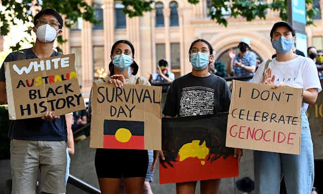 Four people with face masks stand with signs 'white Australia black history', 'Survival Day' and 'I don't celebrate genocide'.