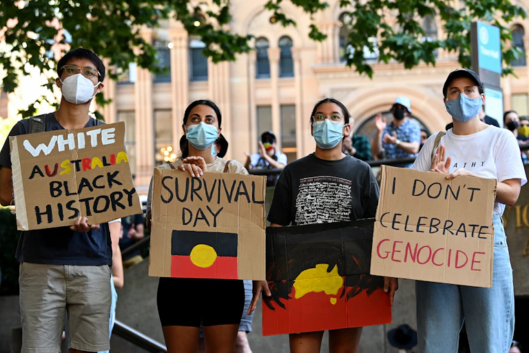 Four people with face masks stand with signs 'white Australia black history', 'Survival Day' and 'I don't celebrate genocide'.