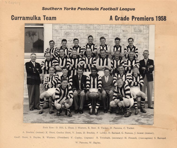 a football team, an Indigenous man smiling in front row, holding a ball