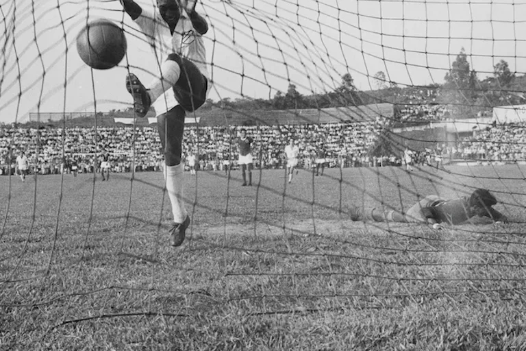 Seorang pemain sepak bola berbaju putih menendang bola ke gawang saat penjaga gawang yang kalah tergeletak di tanah.