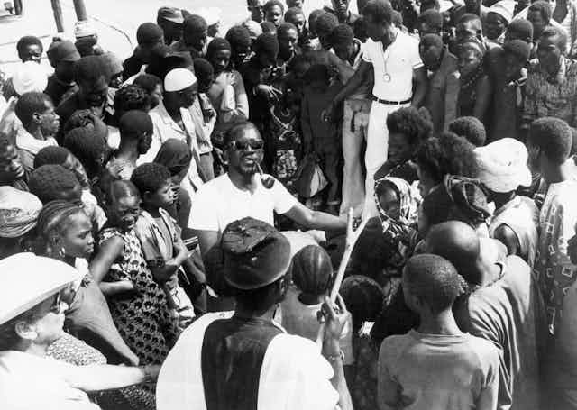 A man in a white T-shirt and shades, a pipe in his mouth, stands in the middle of a large crowd of Africans of all ages.