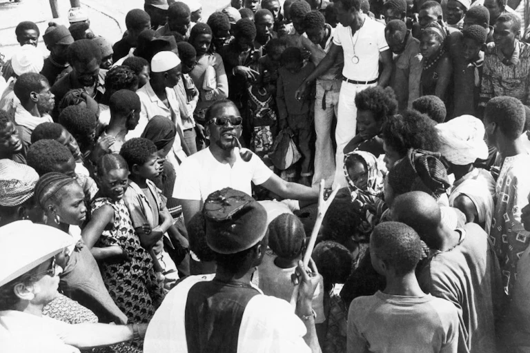 A man in a white T-shirt and shades, a pipe in his mouth, stands in the middle of a large crowd of Africans of all ages.