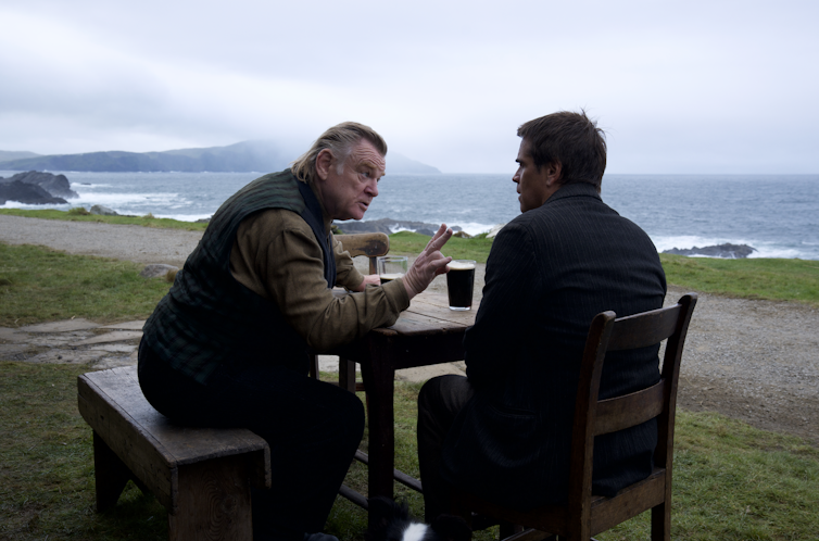 يجلس Brendan Gleeson و Colin Farrell على طاولة حانة في الهواء الطلق مع مكاييل ، مع خلفية من الساحل الأيرلندي.