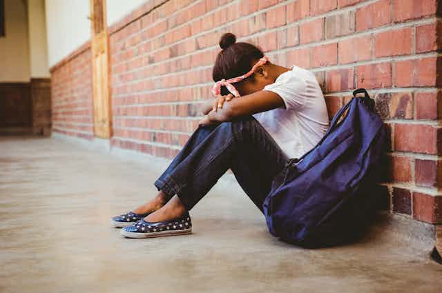 Girl sitting with head on arms next to school bag in corridor