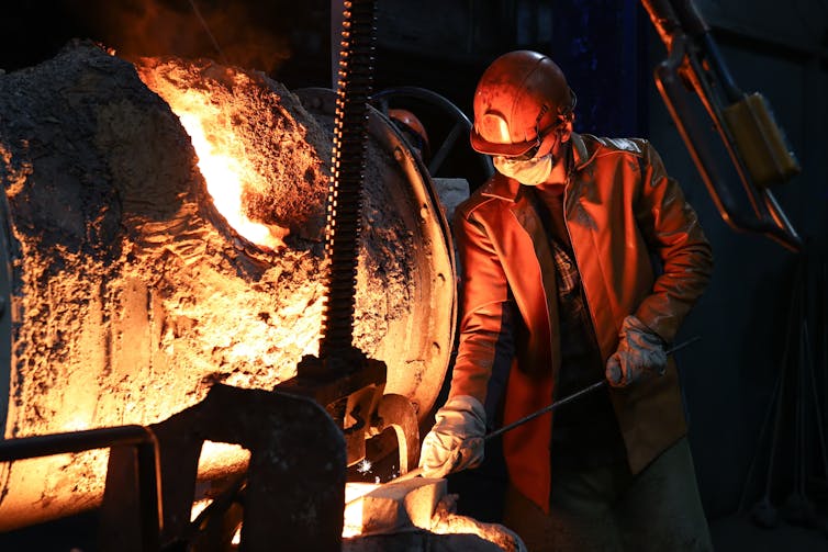 A worker in red overalls pours pig iron from a blast furnace.