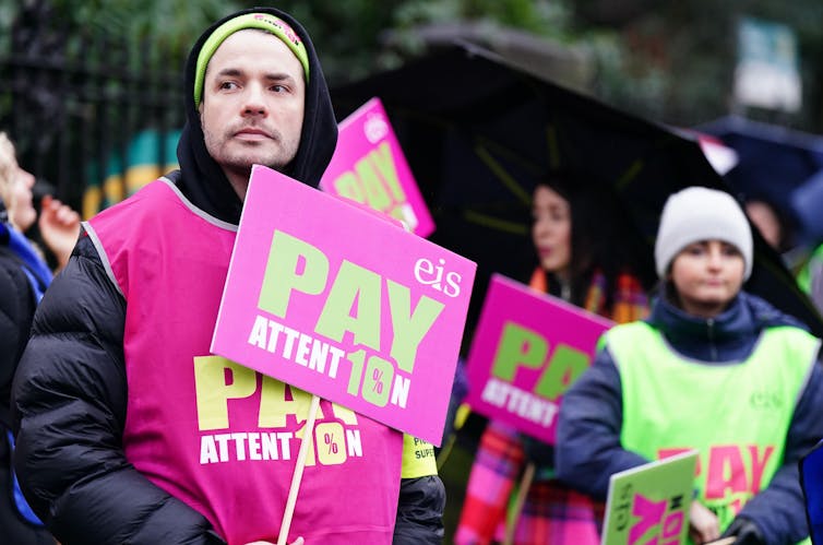 A man holds a sign on a picket line reading 'Pay attention'.