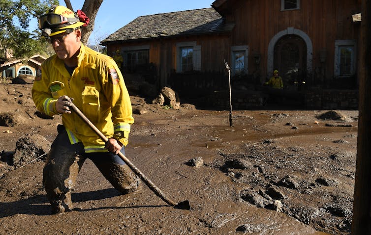 A firefighter knee-deep in mud drags a shovel through the mud in front of a house.