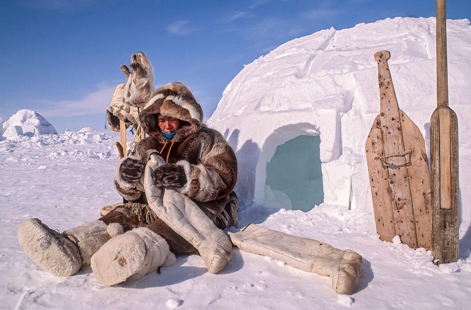 Woman inspects hand sewn boot liners made of caribou skin while sitting outside igloo. She is surrounded by traditional survival tools