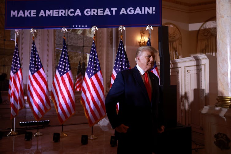 Donald Trump with a blue suit walks past a row of American flags with the words 'Make America Great again' on a banner above the flags.