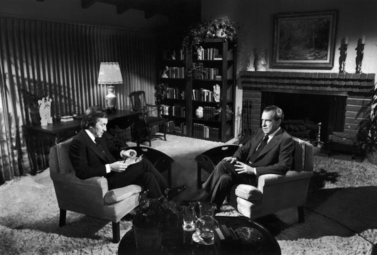 A black and white photo shows two men sitting in arm chairs facing each other.