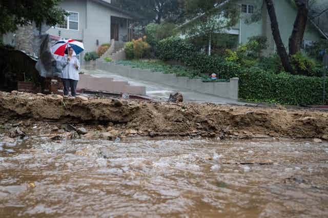 A woman with an umbrella watches a river of mud flow past homes. 