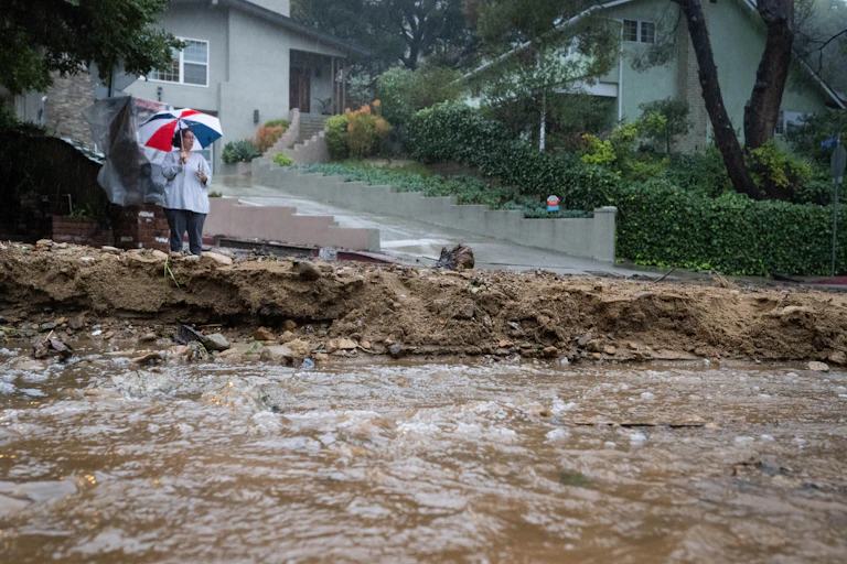 A woman with an umbrella watches a river of mud flow past homes.