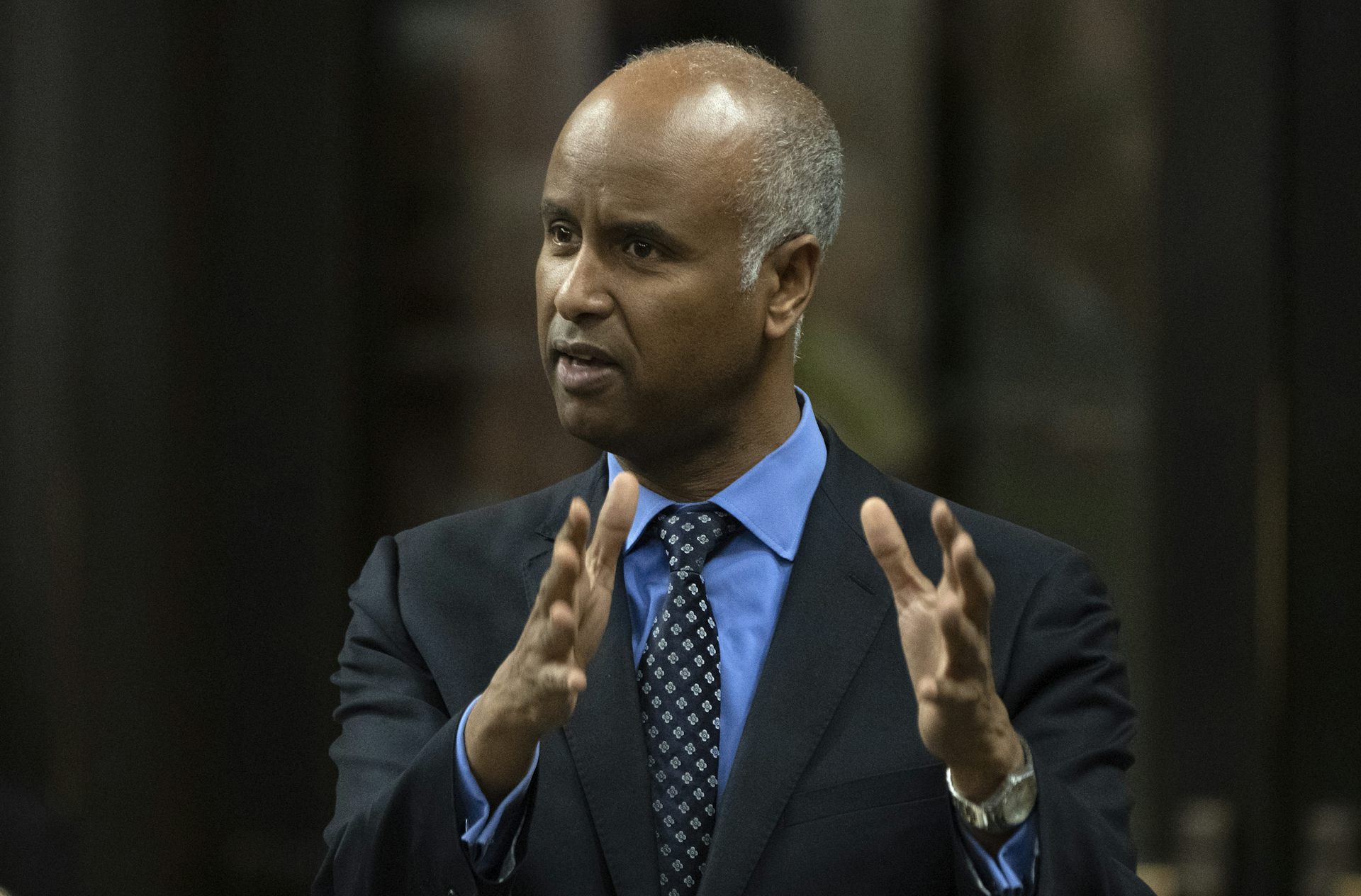 A middle-aged South Asian man in a suit and tie gestures while speaking