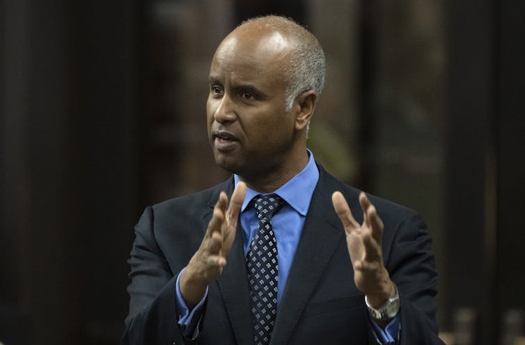 A middle-aged South Asian man in a suit and tie gestures while speaking