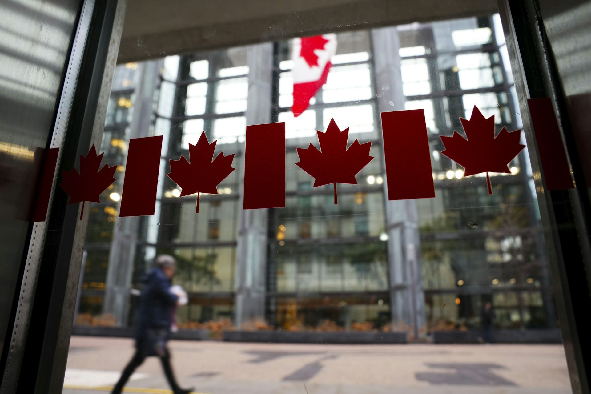 The photo is focused on row of clear Canadian flag sticker in a window. In the background, a person walks past a tall, glass-fronted building.