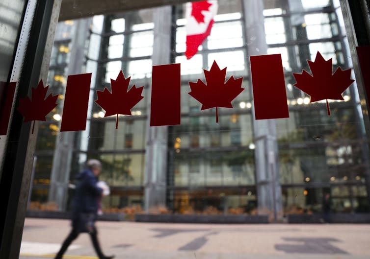 The photo is focused on row of clear Canadian flag sticker in a window. In the background, a person walks past a tall, glass-fronted building.