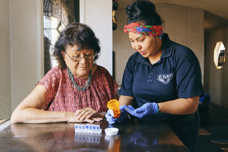 An older Navajo woman receiving health care assistance from someone wearing a shirt emblazoned with the letters E-M-S.