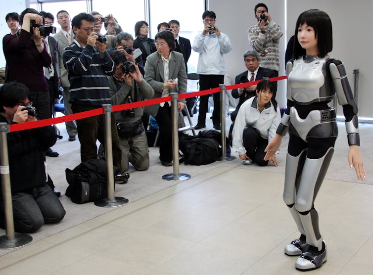A photo of robot that looks like a woman standing in front of a crowd