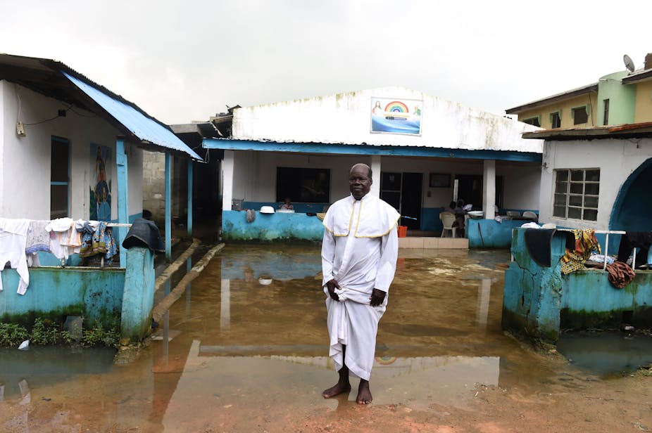 Man wearing a white dress stands in front of a waterlogged building