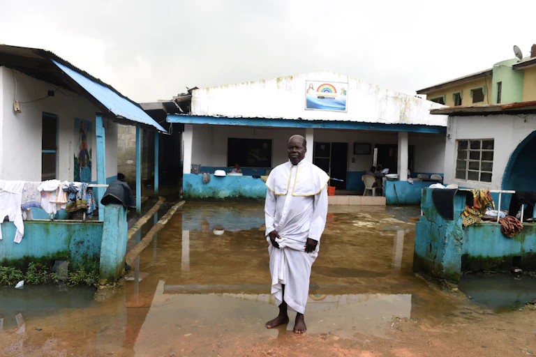 Man wearing a white dress stands in front of a waterlogged building
