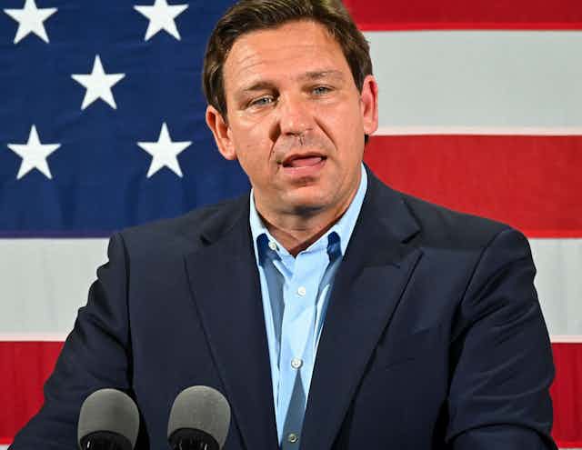 Standing in front of a large American flag, a white man wearing a blue blazer delivers a speech.
