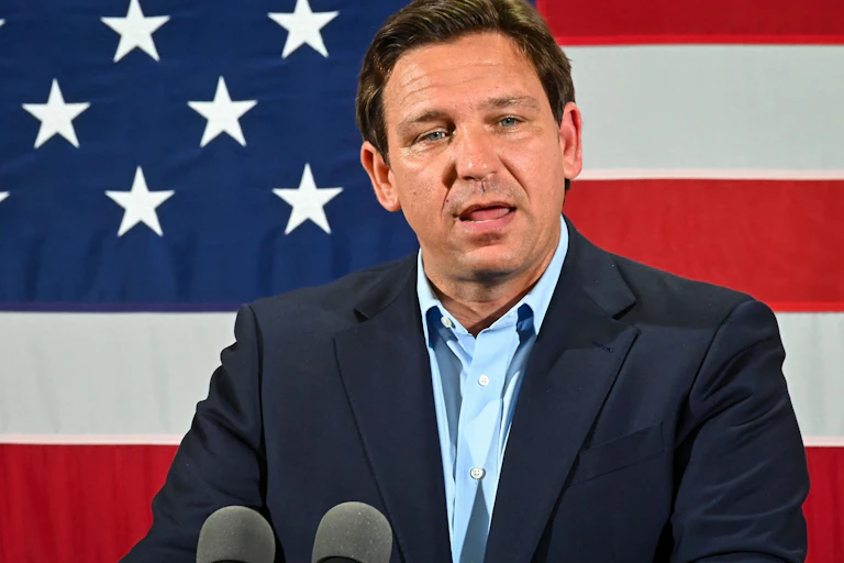 Standing in front of a large American flag, a white man wearing a blue blazer delivers a speech.