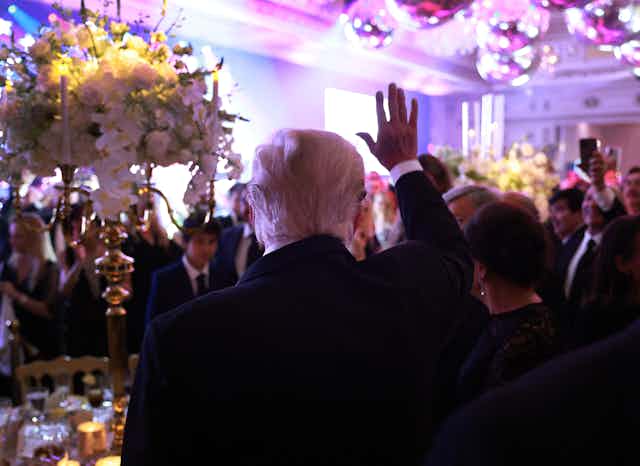 The back of a man with white hair is seen waving his hand, in front of a crowd of people and flowers inside a formal looking room.