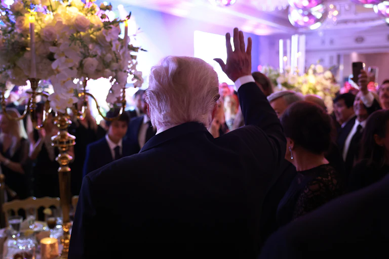 The back of a man with white hair is seen waving his hand, in front of a crowd of people and flowers inside a formal looking room.
