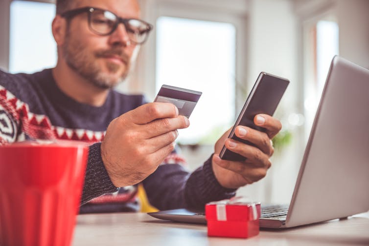 Men wearing Christmas jumper/sweater and eyeglasses holding credit card and using smart phone, laptop, christmas present in foreground.