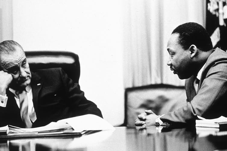A middle-aged white man dressed in a business suit sits on a leather chair and listens to a black man sitting next to him.