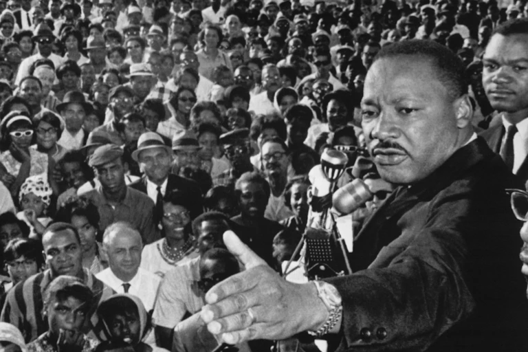 A black man delivers a speech while standing above hundreds of mostly black listeners.