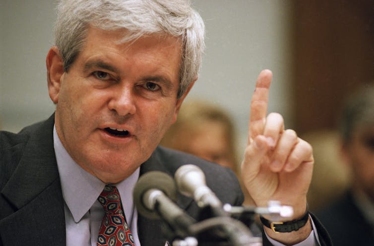 A man in a coat and tie sits in front of a microphone and gestures.