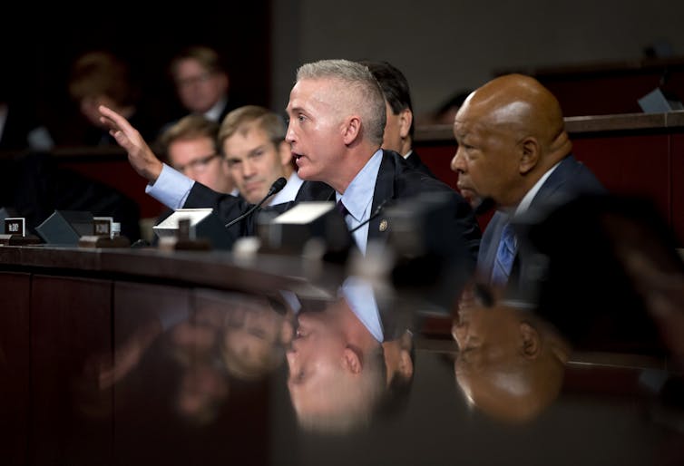 A row of men in suits sit at a table. One leans forward and gestures with his hand.