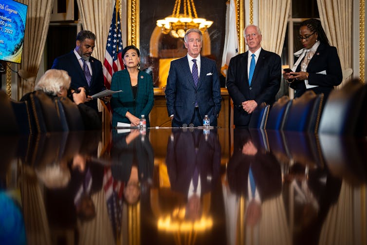 A group of men and women in suits stands around a wooden table.
