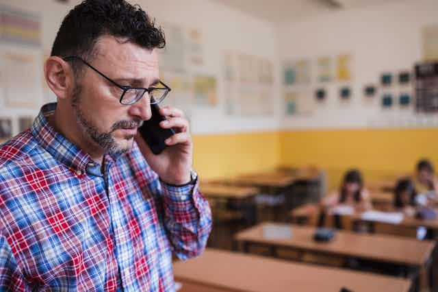 A worried looking teacher talks on a cell phone in class while a few students work at their desks.