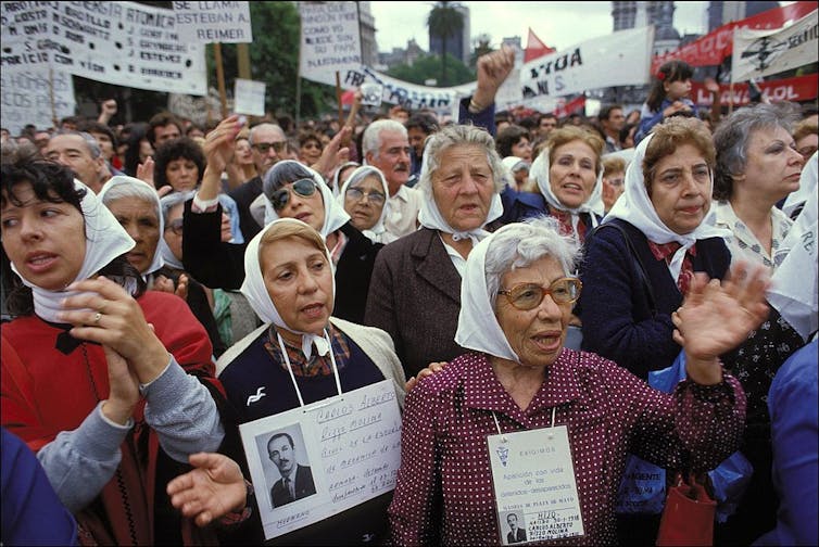 Women wearing white kerchiefs march in a protest, holding placards with photos of people's faces.