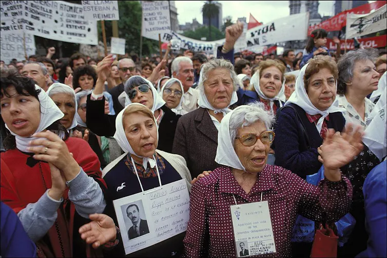 Women wearing white kerchiefs march in a protest, holding placards with photos of people's faces.
