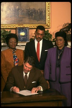 A white man dressed in a business suit is sitting at a desk and signing a piece of paper as two Black women and a young Black man stand behind him