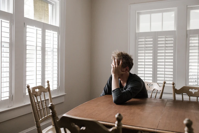 A person looks sadly out the window while obscuring the side of their face with their hand.
