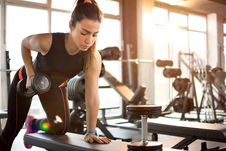 A young woman in gym clothes uses a bench to perform a dumbbell row.