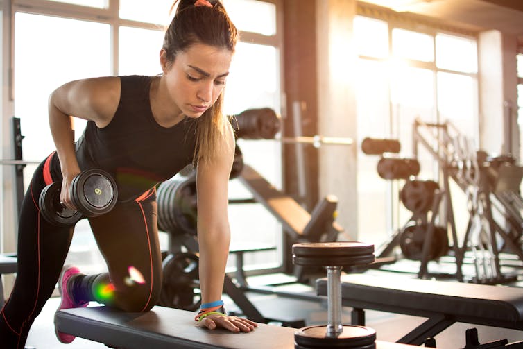 A young woman in gym attire uses a bench to perform a dumbbell row.