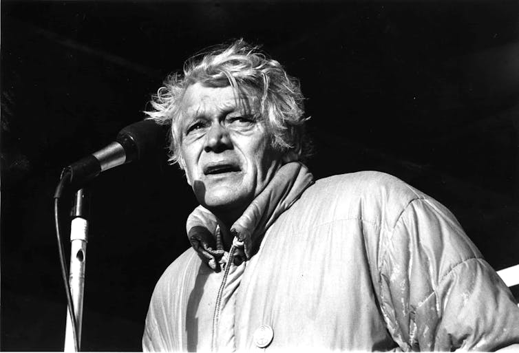 A man dressed in a raincoat stands at a microphone.