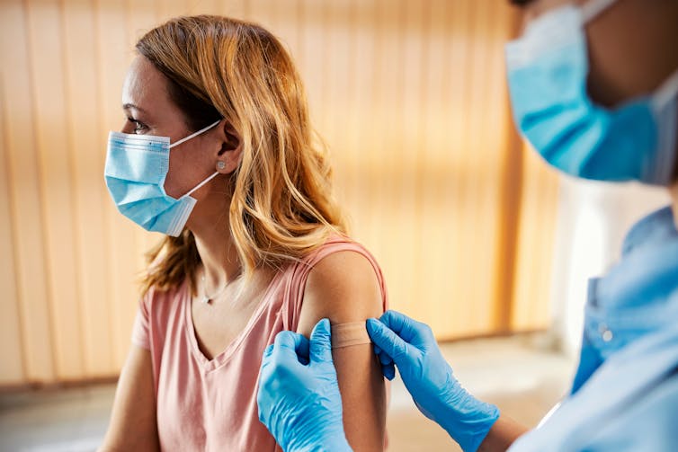 A woman has a bandaid affixed to her upper arm by a healthcare worker.