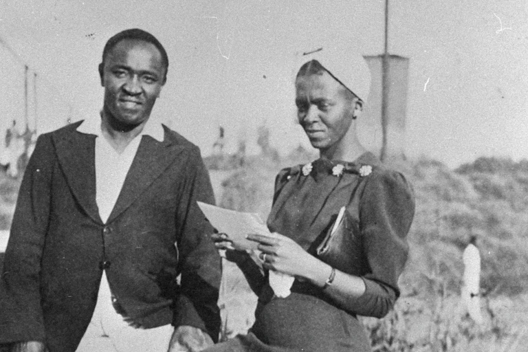 A man and a woman stand posing for photo. The man looks at the camera but the thin, neatly dressed woman looks down at a letter she is holding.