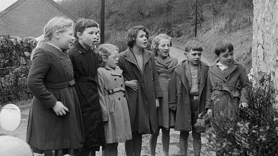A group of children wearing winter coats stand in a line. The year is 1961. 