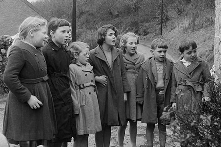 A group of children wearing winter coats stand in a line. The year is 1961.