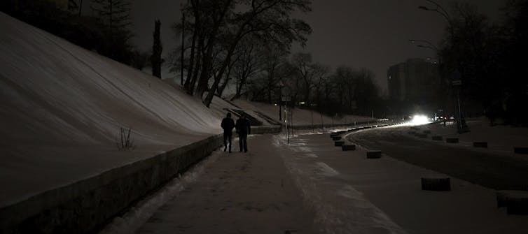 Two people walk along a darkened street with no streetlights at night as a car approaches them.