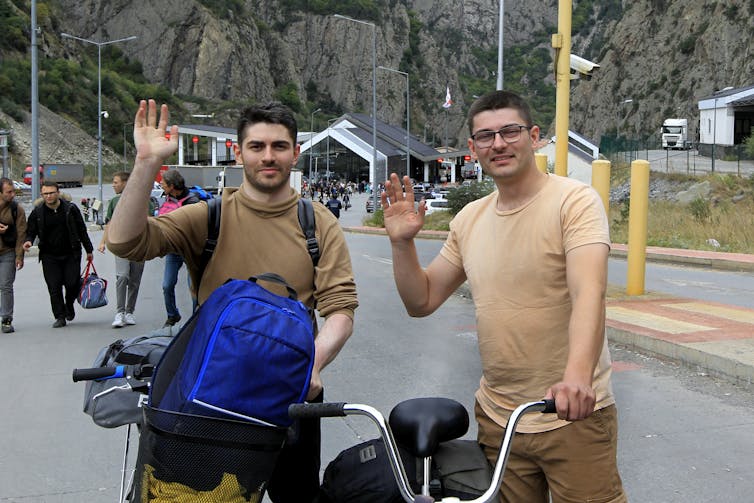 Two men on bicycles wave and smile.