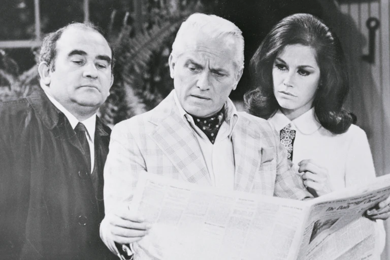 A man wearing an ascot reading a newspaper, flanked by another man and a woman, who are also reading the newspaper.