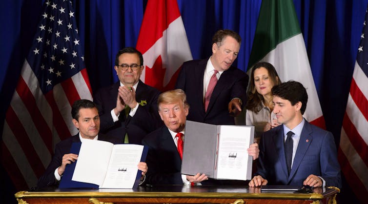 Three men wearing suits sit at a table. Two of them hold up booklets with signatures. Behind them are flags of mexico, canada and the U.S.